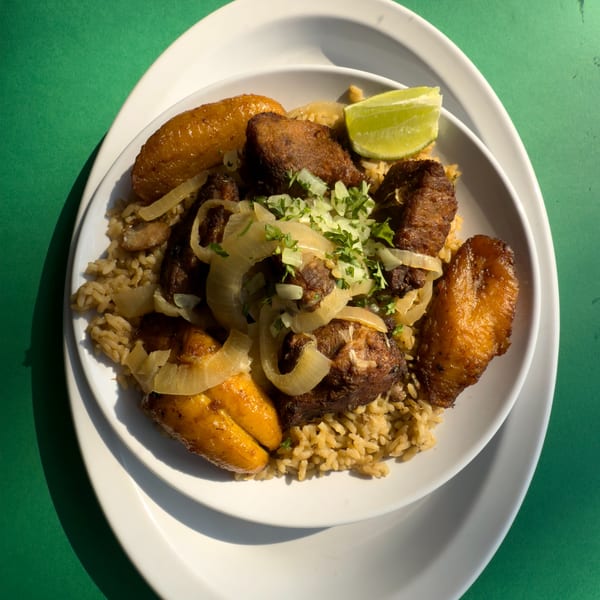Cuban dish with fried pork, rice, plantains, onions, cilantro, and a lime wedge on a white plate.