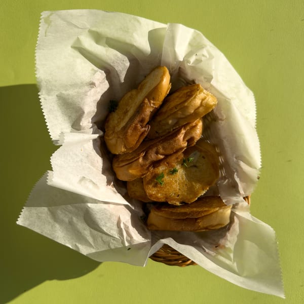 Golden-brown Cuban toast slices, some topped with herbs, in a paper-lined basket on a green table.