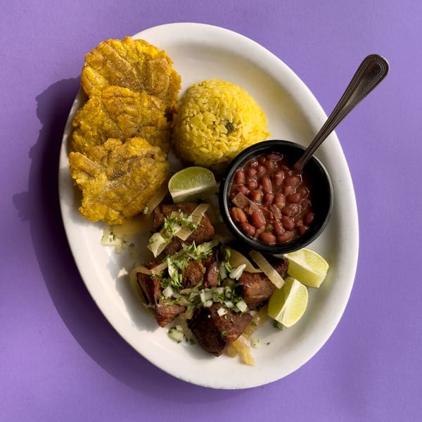 Cuban dish with fried pork, yellow rice, red beans, tostones, and lime wedges on a white plate.