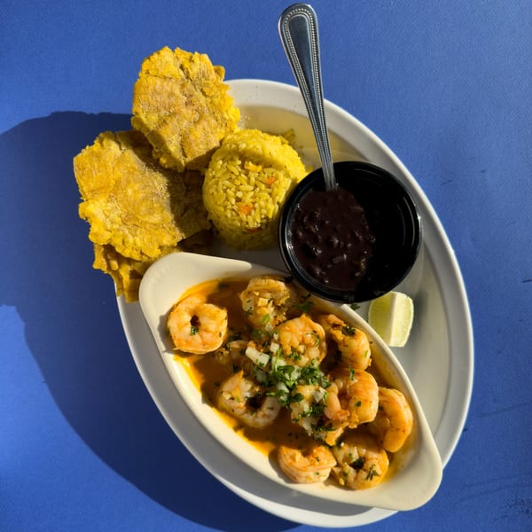 Cuban shrimp creole with yellow rice, black beans, tostones, and a lime wedge on a blue table.