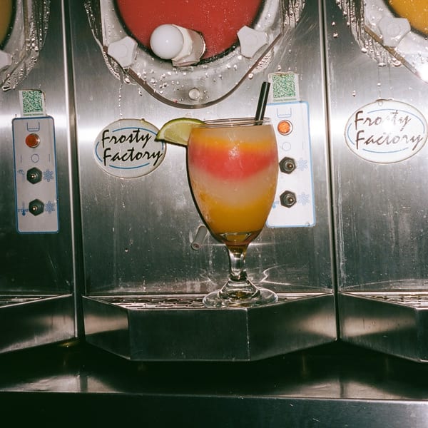 A colorful layered frozen drink with a lime wedge and straw, in front of a "Frosty Factory" machine.