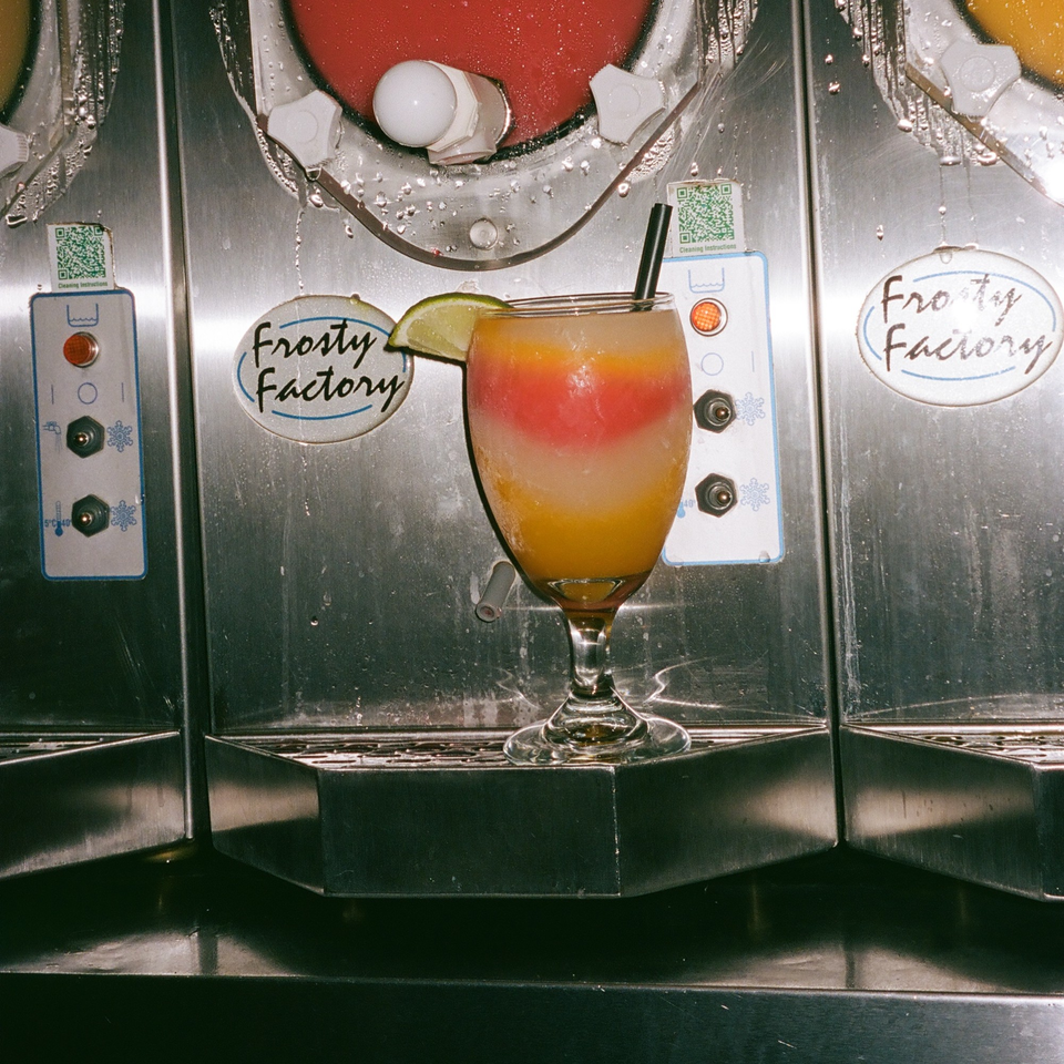 A colorful layered frozen drink with a lime wedge and straw, in front of a "Frosty Factory" machine.