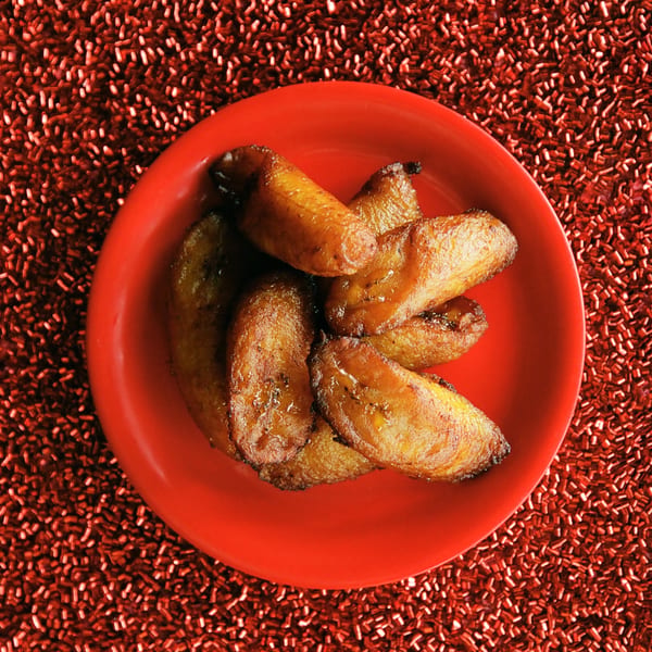 A red plate filled with golden-brown fried plantains, glistening with a sweet glaze, against a red textured background.