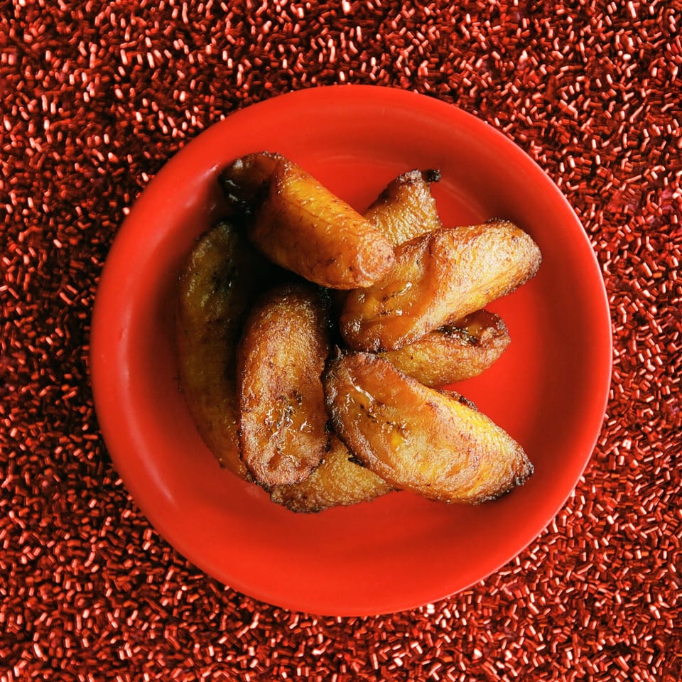 A red plate filled with golden-brown fried plantains, glistening with a sweet glaze, against a red textured background.