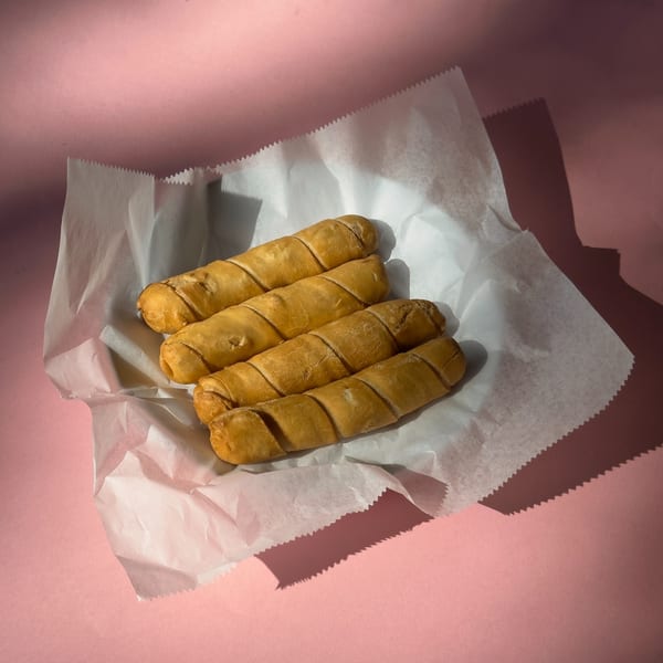 Four golden-brown Cuban tequeños, a fried cheese pastry, rest in a white paper liner on a pink surface.