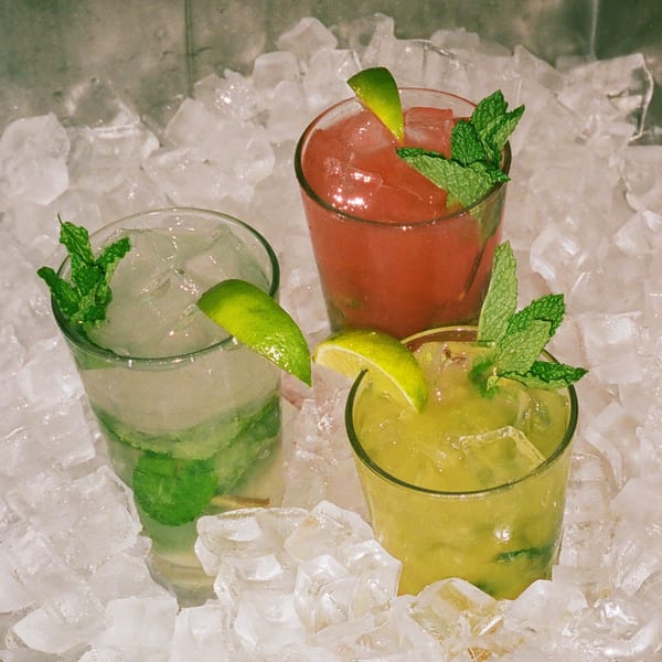 Three vibrant mojitos, red, clear, and green, garnished with lime and mint, nestled in ice.